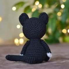 a crocheted black teddy bear sitting in front of a christmas tree with lights