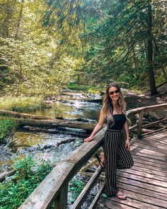 a woman standing on a wooden bridge over a stream in the woods with trees and water behind her