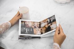 a person holding an open photo book on top of a white table next to a cup