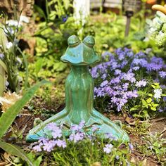 a green frog statue sitting in the middle of some purple and white flowers on the ground