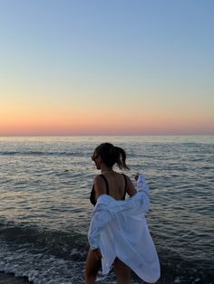 a woman in a white dress walking along the beach at sunset with her back to the camera