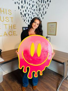 a woman holding up a large pink and yellow smiley face sign in front of a desk