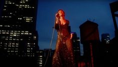 a woman standing in front of a microphone on top of a roof with buildings behind her