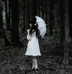 a woman in a white dress holding an umbrella standing in the woods with leaves on the ground