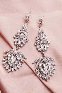 two pairs of earrings on top of a pink satin material surface, one with crystal drops and the other with white stones