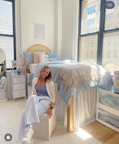 a woman sitting on a bed in a room with large windows and white walls, smiling at the camera