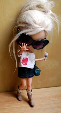 a doll with blonde hair wearing sunglasses and holding a small object in her hand while standing next to a wall