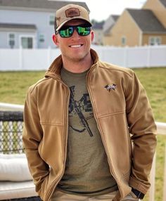 a man wearing sunglasses and a jacket standing in front of a fence with his hands on his hips