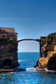 an arch in the rock with a bridge above it on top of blue ocean water