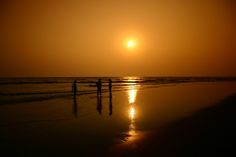 two people walking on the beach at sunset with the sun setting in the distance behind them