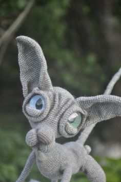 a knitted toy rabbit with blue eyes and ears, standing in front of some trees