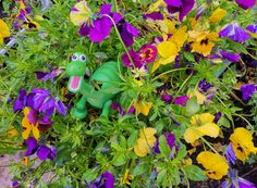 a green toy sitting in the middle of some purple and yellow flowers