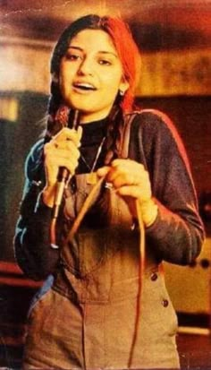 a woman holding a microphone in her right hand and wearing overalls on the other side