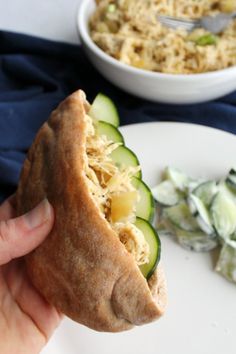 a hand holding a sandwich with cucumbers and noodles in the background