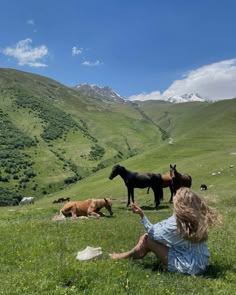 a woman sitting in the grass next to horses and cows on a hill side with mountains in the background