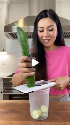 a woman holding up a cucumber in front of a cup on top of a counter