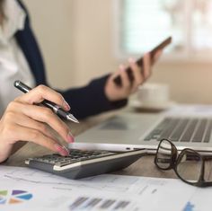 a woman sitting at a desk using a calculator and cell phone to work on her laptop