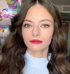 a woman with long brown hair wearing a white turtle neck sweater and pink lipstick looks at the camera