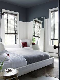 a white bed sitting next to two windows with shutters on each side and a table in front of it