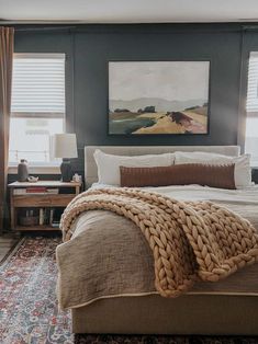 a large bed sitting in a bedroom next to two windows with blinds on them and a painting hanging above it