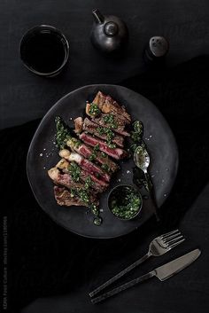 a plate with steak and greens on it next to two silver spoons, cups and saucers