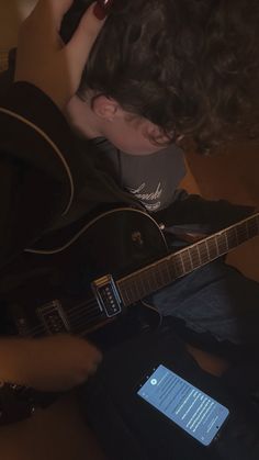 a young man is playing the guitar and looking at his cell phone while sitting down