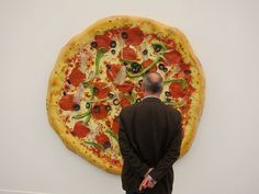 a man standing in front of a large pizza on display at an art gallery with his back to the camera