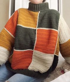 a woman wearing a multicolored crocheted sweater sits in front of a window