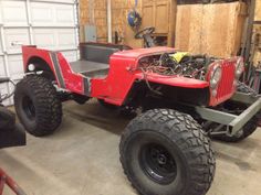 an old red jeep is parked in a garage with large tires on it's flatbed