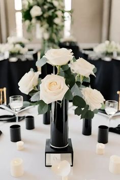 a black vase with white flowers on top of a table surrounded by candles and place settings