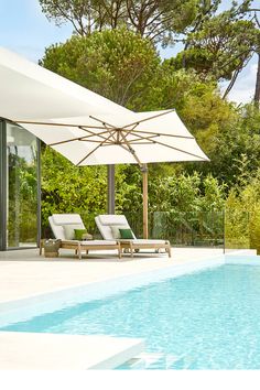 two lounge chairs under an umbrella next to a swimming pool with trees in the background