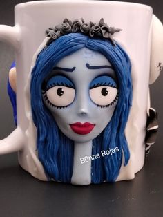 a white coffee mug with blue hair and makeup painted on it's face is sitting on a table