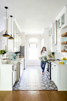 a woman standing in the middle of a kitchen with white cabinets and wood flooring