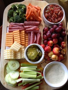 a wooden tray topped with lots of different types of cheeses and vegetables next to dip