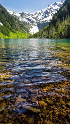 a mountain lake surrounded by green trees and rocks in the foreground with snow capped mountains in the background