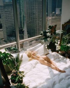 a woman laying on top of a white bed in a bedroom next to tall buildings