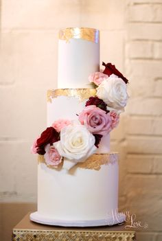 a white and gold wedding cake with flowers on it's tiered stand, against a brick wall