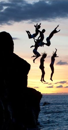 some people jumping into the air on top of a cliff by the ocean at sunset