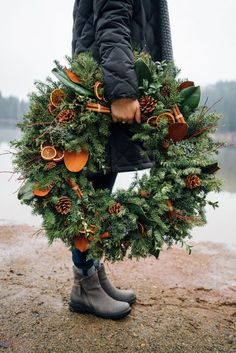 a person holding a wreath with pine cones and oranges on it in front of a body of water