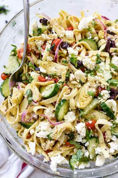 a glass bowl filled with pasta salad on top of a white table next to utensils