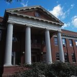 an old brick building with columns and a clock on the top floor is surrounded by greenery