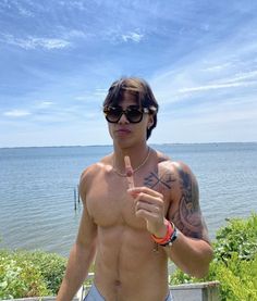 a shirtless man with sunglasses on holding a finger up to the camera while standing in front of a body of water