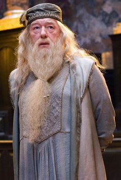 an old man with long white hair and beard wearing a gray outfit standing in front of a table