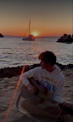 a young man sitting on the beach playing an acoustic guitar as the sun sets in the background