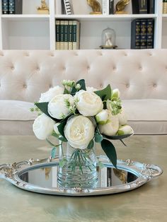 a vase with white flowers on a tray in front of a couch and bookshelf