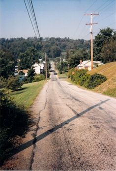 an empty dirt road with power lines above it and houses in the distance on either side