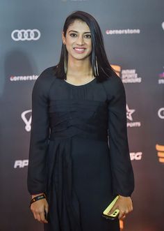 a woman standing on a red carpet wearing a black dress and gold jewelry, smiling at the camera