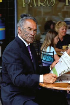 a man in a suit and tie sitting at a table reading a paper with tattoos on his face