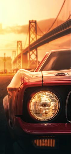 the front end of a red sports car in front of a suspension bridge at sunset