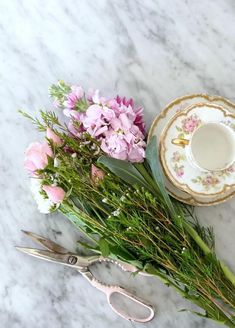 flowers and scissors on a marble table with a teacup in the middle, next to it is a pair of scissors